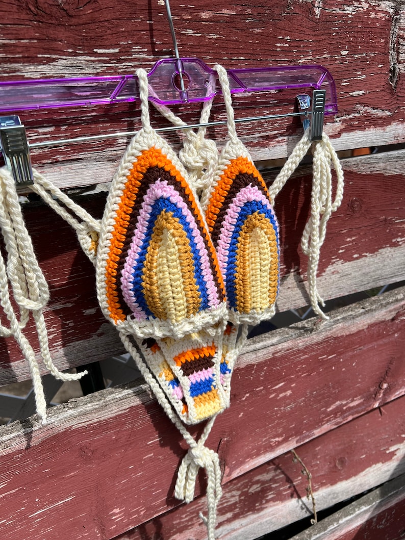 May include: A crocheted bikini top and bottom with a rainbow pattern in shades of orange, brown, pink, blue, and yellow. The bikini is made of white yarn and has a tie closure.