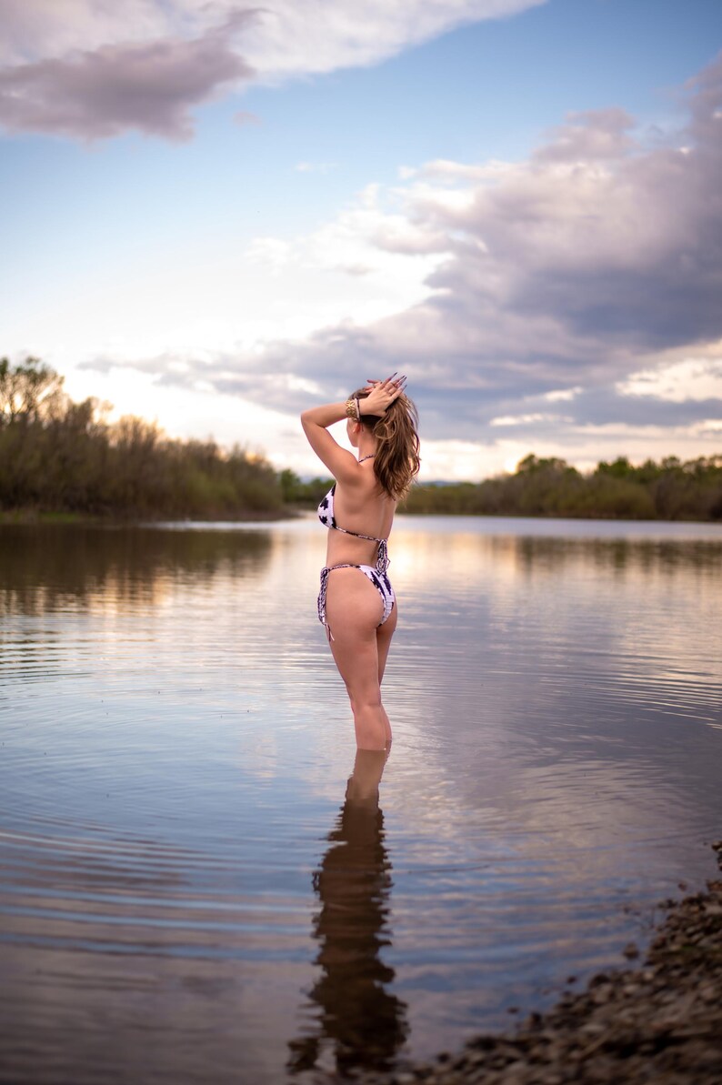 May include: A two-piece bikini featuring a black and white cow print pattern. The top is a triangle style with thin straps, and the bottom is a high-cut design with side ties. The swimwear is shown from a rear view, highlighting the pattern.