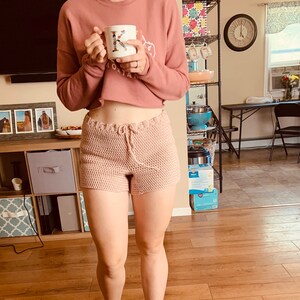 May include: A woman wearing a pink cropped sweater and pink crocheted shorts. She is holding a white mug with a floral design and the letter K on it.