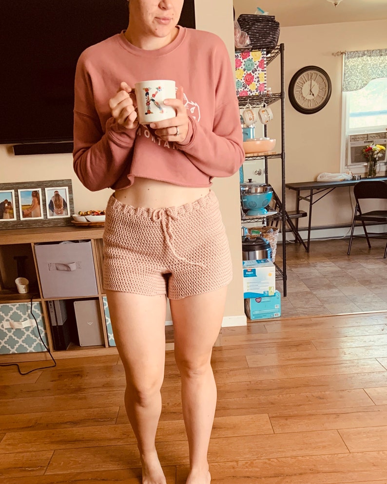May include: A woman wearing a pink cropped sweater and pink crocheted shorts. She is holding a white mug with a floral design and the letter K on it.