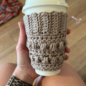 May include: A beige crocheted coffee cup cozy with a textured pattern. The cozy is on a white to-go cup.