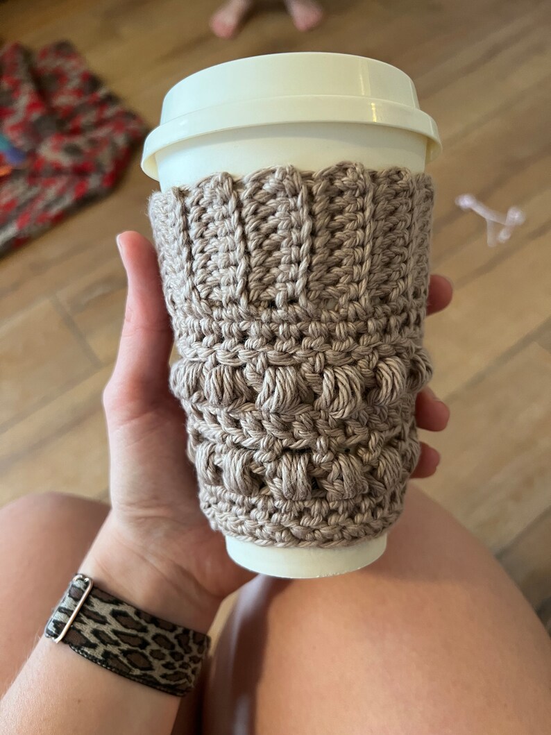 May include: A beige crocheted coffee cup cozy with a textured pattern. The cozy is on a white to-go cup.