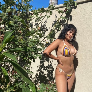 May include: A woman wearing a colorful crocheted bikini top and matching bottom. The bikini is a rainbow of colors, including orange, yellow, red, purple, and blue. The woman is standing in front of a white wall with a green bush behind her.