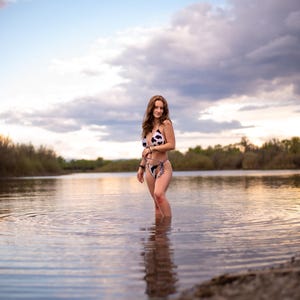 May include: A two-piece bikini featuring a black and white cow print pattern. The top is a triangle style with thin straps, and the bottom is a high-cut design with side ties. The swimwear is worn in a natural outdoor water setting.