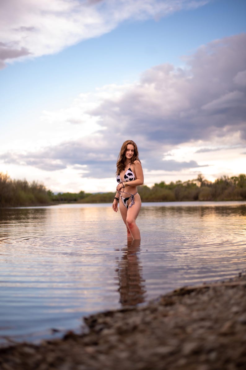 May include: A two-piece bikini featuring a black and white cow print pattern. The top is a triangle style with thin straps, and the bottom is a high-cut design with side ties. The swimwear is worn in a natural outdoor water setting.
