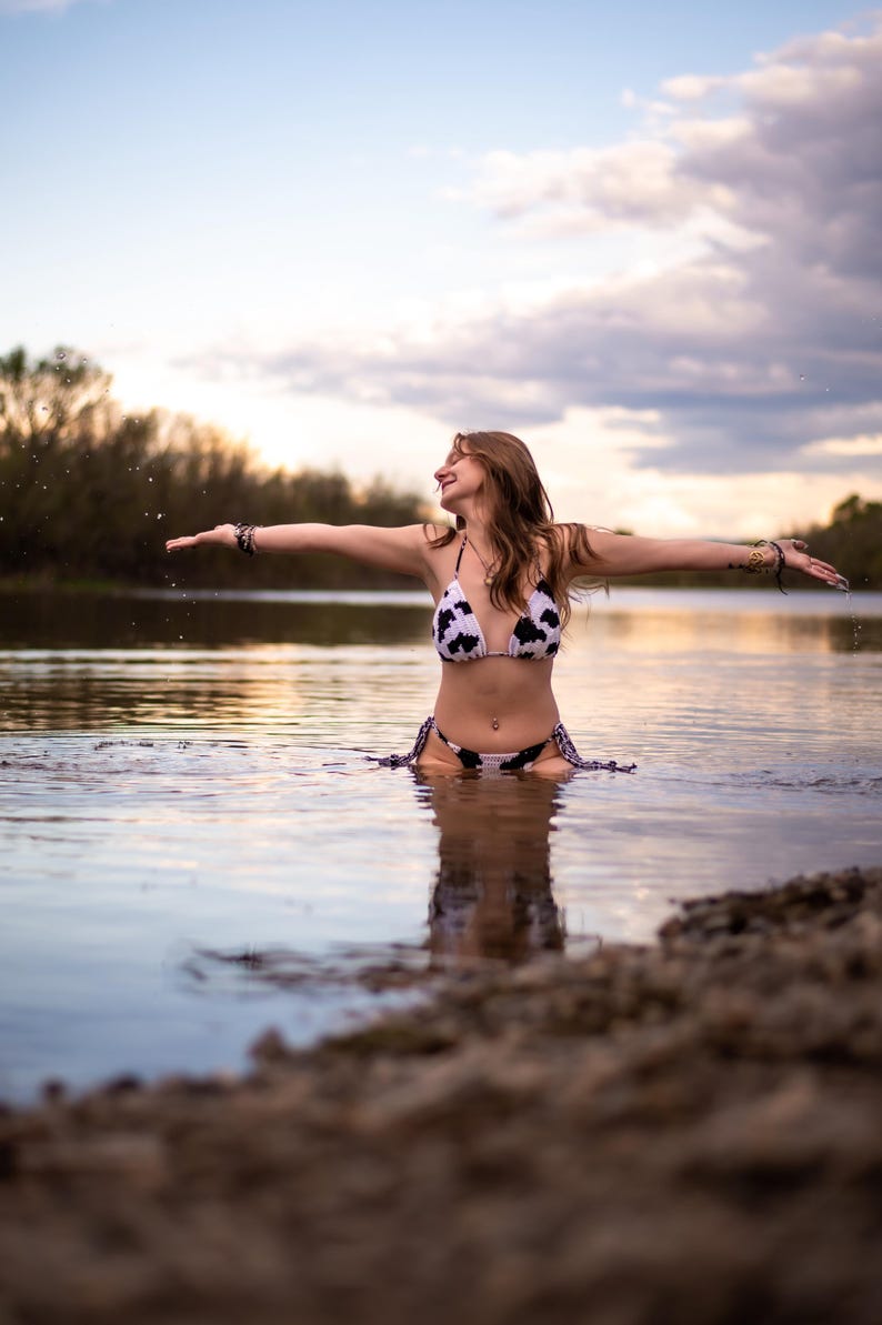 May include: A two-piece bikini featuring a black and white cow print pattern. The top is a triangle style with thin straps, and the bottom is a side-tie style. The swimwear is constructed from a fabric material and is worn in a body of water.