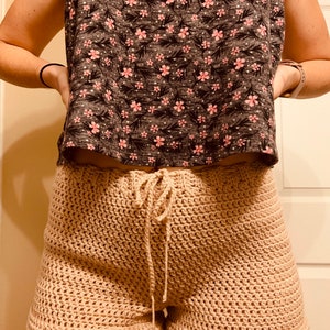 May include: Beige crochet shorts with a drawstring waist. The shorts are made with a simple stitch pattern and have a relaxed fit.