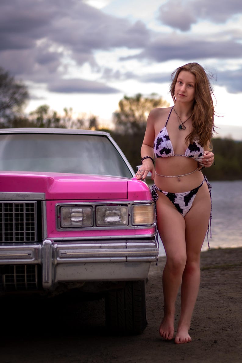 May include: A two-piece crochet bikini featuring a white and black cow print pattern with side ties, worn with a thin silver-toned belly chain and a dark pendant necklace, standing beside the front corner of a bright pink vintage car.