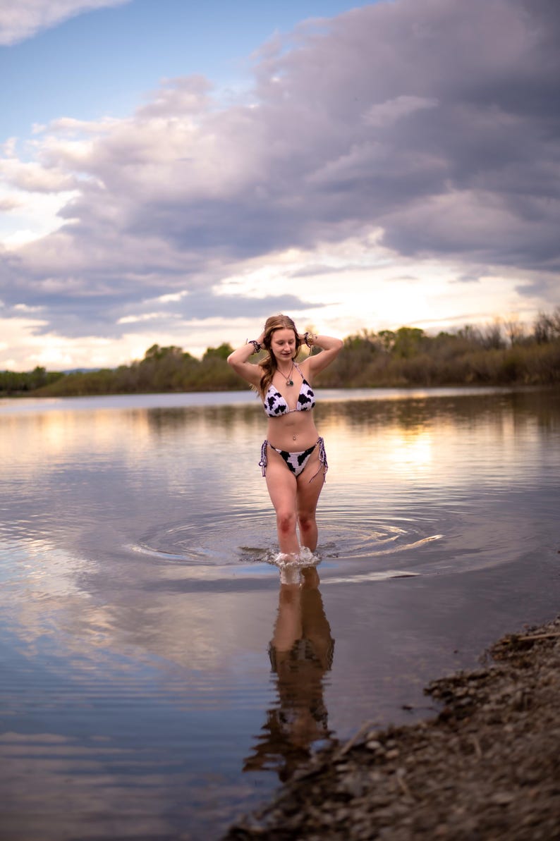 May include: A two-piece bikini set featuring a black and white cow print pattern. The top is a triangle style with thin straps, and the bottom is a classic cut with side ties. The swimwear is made of a smooth fabric with a consistent spotted design.