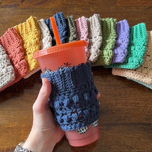 May include: A collection of crocheted cup cozies in various colors, including beige, orange, yellow, green, purple, blue, and light blue. The cozies are made with a textured stitch and have a raised, bumpy design. One of the cozies is on a pink cup with a straw.