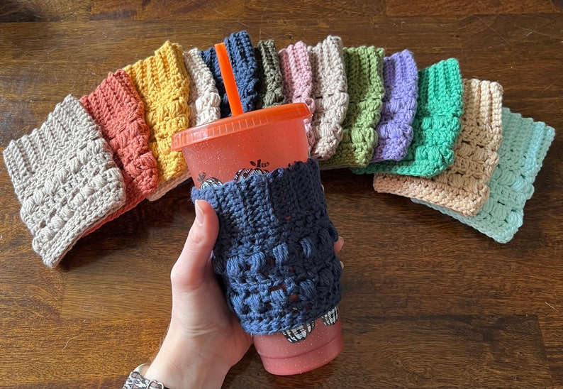 May include: A collection of crocheted cup cozies in various colors, including beige, orange, yellow, green, purple, blue, and light blue. The cozies are made with a textured stitch and have a raised, bumpy design. One of the cozies is on a pink cup with a straw.