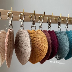 May include: A set of seven colorful tassel keychains hanging from a wooden rack. The keychains are made of yarn and have a fringe-like design. The colors of the keychains are beige, mustard yellow, burgundy, teal, and blue.