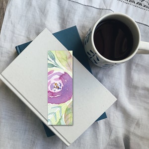 Pode incluir: Um marcador de p&aacute;gina de aquarela com uma flor roxa e folhas verdes em um fundo verde claro.