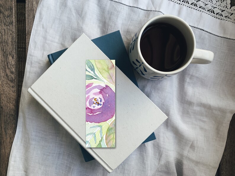 Pode incluir: Um marcador de p&aacute;gina de aquarela com uma flor roxa e folhas verdes em um fundo verde claro.