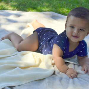May include: A baby wearing a blue and white patterned onesie lies on a white blanket on the grass. The baby is looking at the camera and smiling.