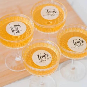 May include: Four champagne glasses filled with a golden liquid, each with a sticker reading "Team Bride". The glasses have a textured design and sit on a light-coloured surface. A celebratory theme is evident.