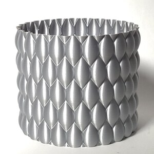 May include: A silver-coloured, cylindrical planter with a textured, geometric pattern resembling scales. The planter is made of a smooth, shiny material.
