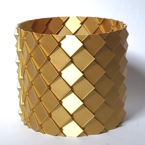 May include: A gold-colored, geometric, cylindrical container made of interlocking diamond-shaped pieces. The container has a light yellow stripe running vertically down the side.