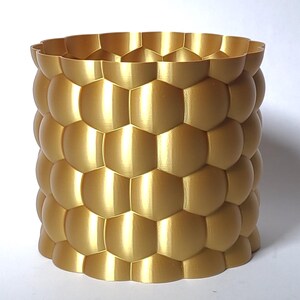 May include: A gold-coloured cylindrical planter with a textured surface. The planter is made of a 3D printed material and has a honeycomb-like pattern.