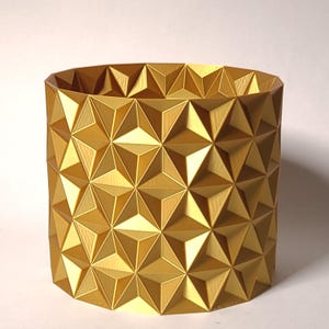 May include: A gold-colored cylindrical planter with a geometric pattern of triangles. The planter is made of a smooth, shiny material.