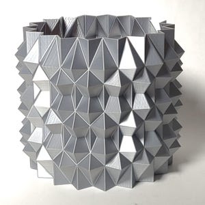 Puede incluir: Un macetero geométrico plateado con un patrón texturizado en forma de diamante. El macetero está hecho de un material duradero y tiene un diseño moderno.