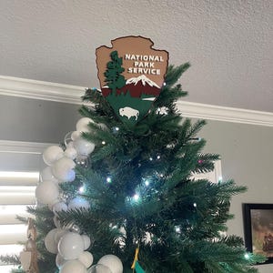 May include: A Christmas tree topped with a National Park Service emblem. The emblem is a brown, green, and white shield shape with the words "NATIONAL PARK SERVICE" and a mountain, tree, and bison illustration. The tree is decorated with white ornaments and lights.