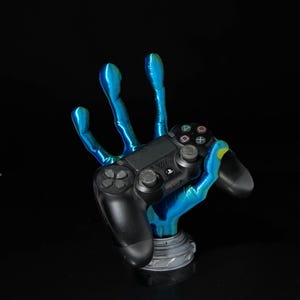 May include: A blue and black gaming controller rests in a blue, hand-shaped stand. The stand has a metallic base and a vibrant, iridescent finish. The controller is black with buttons and directional controls.