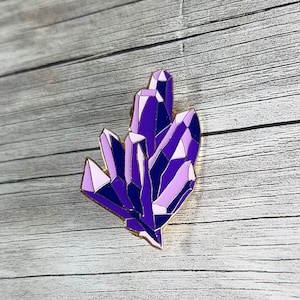 May include: A purple and pink crystal cluster enamel pin with a gold outline. The pin is a geometric design with sharp edges and points.