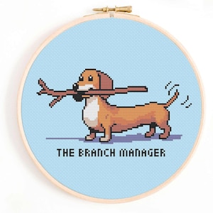 Könnte beinhalten: Ein Kreuzstich-Stickrahmen mit hellblauem Hintergrund, auf dem ein Cartoon-Dackel zu sehen ist, der einen Stock im Maul trägt. Unter dem Dackel steht der Text "THE BRANCH MANAGER".