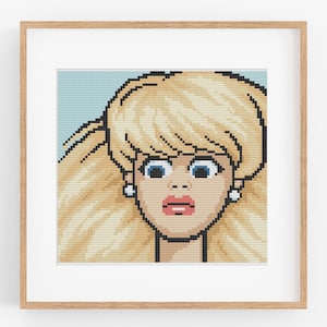 May include: A framed pixel art print depicting a blonde woman with blue eyes and a surprised expression. The artwork is in a cross-stitch style, set against a light blue backdrop. The frame is made of light brown wood.