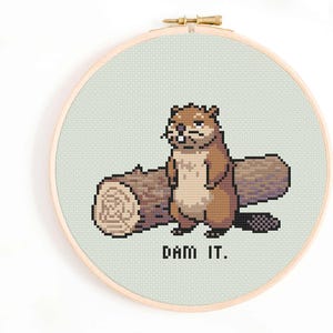 May include: Cross-stitch embroidery featuring a brown beaver and a log, set against a pale green backdrop. The text "DAM IT." is stitched below the beaver. The embroidery is framed in a wooden hoop.
