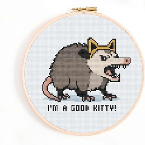 Op de afbeelding: Kruissteekborduurwerk in een houten hoepel met een gepixeleerde opossum met een kroon en ontblote tanden. De tekst "I'M A GOOD KITTY!" staat eronder. De achtergrond is lichtblauw.