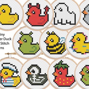 May include: A collection of twelve tiny rubber duck cross-stitch patterns, each displayed in a small wooden embroidery hoop. The ducks are various colors and designs, including a yellow duck, a red devil duck, and a bee duck. The text "12 x Tiny Rubber Duck Cross Stitch Patterns" is visible.