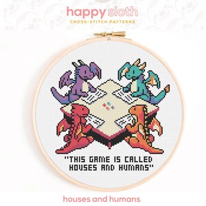 May include: A cross-stitch pattern in a wooden hoop shows four pixelated dragons playing a game. The dragons are purple, teal, red, and orange. The text "THIS GAME IS CALLED HOUSES AND HUMANS" is below the game scene. The pattern is titled "houses and humans".