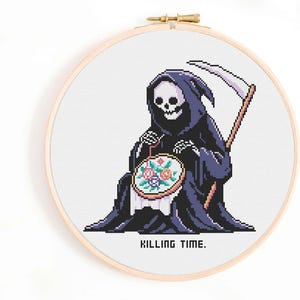 May include: Cross-stitch embroidery hoop featuring a pixelated Grim Reaper in a black cloak, holding a floral embroidery. The text "KILLING TIME" is below the image. The hoop is set against a white background.