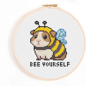 May include: Cross-stitch embroidery in a wooden hoop features a guinea pig dressed as a bee. The bee costume is yellow and black striped, with blue wings. The text "BEE YOURSELF" is stitched below the guinea pig.