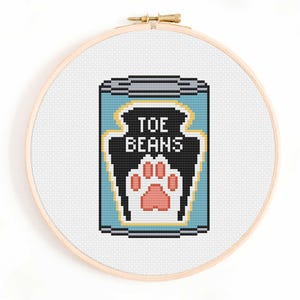 May include: A cross-stitch embroidery in a round wooden hoop featuring a light blue can with a black label. The label displays the white text TOE BEANS above a pink and white paw print graphic. The can has a grey lid and base with yellow trim.
