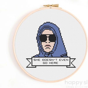 May include: Cross-stitch pattern featuring a pixelated illustration of a person wearing a blue hoodie and sunglasses with the text "She doesn't even go here" on a banner.