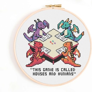 Puede incluir: Bordado de punto de cruz en un aro de madera. El diseño presenta cuatro dragones pixelados de diferentes colores jugando a un juego alrededor de una mesa. El texto dice "THIS GAME IS CALLED HOUSES AND HUMANS".
