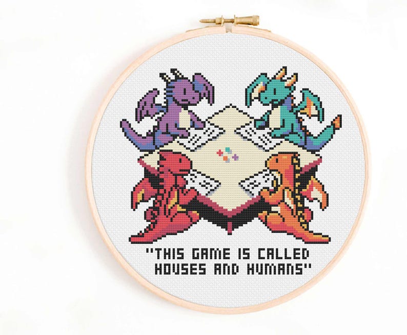May include: A cross-stitch embroidery in a wooden hoop depicts four pixelated dragons playing a game. The dragons are purple, green, red, and orange, gathered around a table. The text "THIS GAME IS CALLED HOUSES AND HUMANS" is below the table.