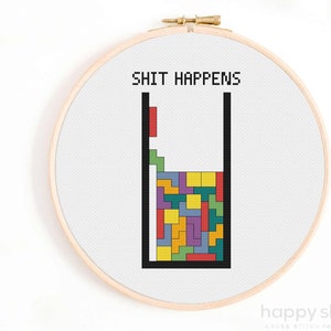 以下が含まれることがあります： 「SHIT HAPPENS」というテキストと、進行中のテトリスゲームのピクセル化された画像が描かれたクロスステッチパターン。ゲームは黒い輪郭のあるグラスの中にあります。ブロックは赤、オレンジ、黄、緑、青、紫、黒です。