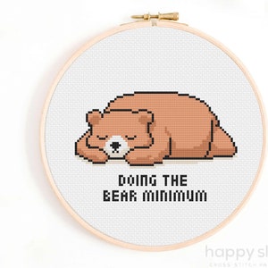 Puede incluir: Un bastidor de punto de cruz con un oso pardo durmiendo y el texto "DOING THE BEAR MINIMUM". El oso está en un estilo pixelado, y el texto está en letras negras. El bastidor es de madera.