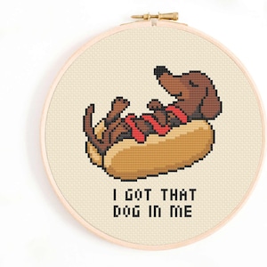 May include: Cross-stitch embroidery in a wooden hoop. The design shows a brown dachshund in a hot dog bun, wearing a red collar. The text "I GOT THAT DOG IN ME" is below the image.