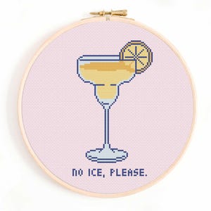 Op de afbeelding: Kruissteekborduurwerk in een houten hoepel met een gepixelde margarita-cocktail met een schijfje citroen. Het ontwerp staat tegen een lichtroze achtergrond, met de tekst "NO ICE, PLEASE." onderaan.
