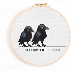 May include: Cross-stitch embroidery featuring two pixelated black crows with orange eyes. The phrase "ATTEMPTED MURDER" is stitched below the birds. The artwork is displayed in a wooden embroidery hoop.