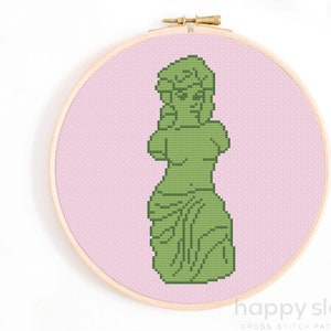 May include: A cross stitch pattern of a green statue of a woman on a pink background. The pattern is in a wooden embroidery hoop.