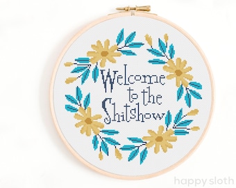 以下が含まれることがあります： 花輪と「Welcome to the Shitshow」というテキストが書かれたクロスステッチパターンです。