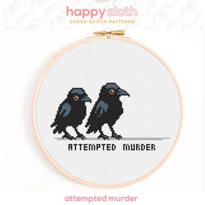 Puede incluir: Patr&oacute;n de punto de cruz con dos cuervos negros con ojos naranjas, uno al lado del otro. La frase "ATTEMPTED MURDER" est&aacute; debajo de los p&aacute;jaros. El dise&ntilde;o est&aacute; en un bastidor de bordado de madera, con el logotipo de Happy Sloth en la parte superior.