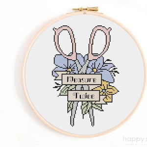 Op de afbeelding: Kruissteekborduurwerk in een houten hoepel met een roze schaar, blauwe en gele bloemen en de tekst "Measure Twice". Het ontwerp bevat groene bladeren en een banner. De woorden "happy sloth" staan onderaan.