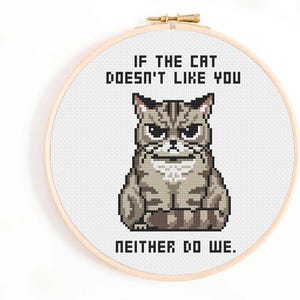 May include: Cross-stitch embroidery in a wooden hoop features a grumpy grey cat with the text "IF THE CAT DOESN'T LIKE YOU NEITHER DO WE." The cat is pixelated, with a stern expression.